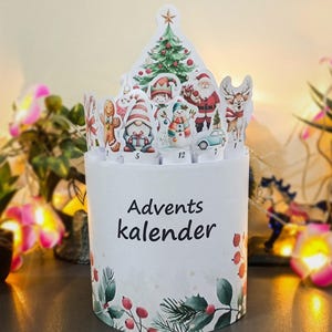 Puede incluir: Un calendario de Adviento blanco con un diseño festivo. El contenedor cilíndrico presenta ilustraciones de personajes navideños y un árbol de Navidad. El texto "Advents kalender" está impreso en la parte delantera. La base está decorada con acebo y bayas.