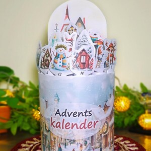 Peut inclure: Un calendrier de l'Avent cylindrique avec une scène de village d'hiver et l'inscription "Advents kalender". Le haut présente des découpes de personnages festifs comme un bonhomme de neige, des maisons et un renne. Le calendrier est posé sur un dessous de plat rouge et beige.