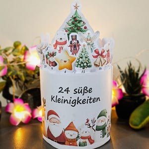 May include: A white Christmas advent calendar with 24 compartments, featuring festive illustrations of Santa Claus, snowmen, and a star. The top has cut-out shapes of Christmas trees and other holiday figures. The text "24 s&uuml;&szlig;e Kleinigkeiten" is printed on the front.