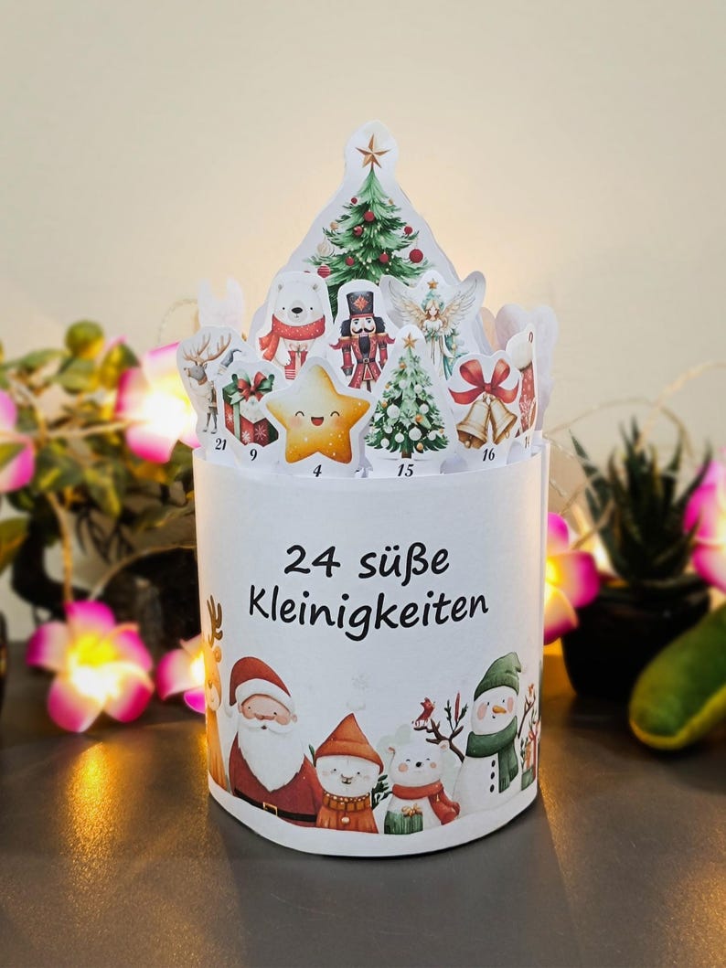 May include: A white Christmas advent calendar with 24 compartments, featuring festive illustrations of Santa Claus, snowmen, and a star. The top has cut-out shapes of Christmas trees and other holiday figures. The text "24 s&uuml;&szlig;e Kleinigkeiten" is printed on the front.