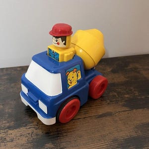 May include: A blue and white toy cement mixer truck with red wheels and a yellow mixer. A yellow and red figure sits in the driver's seat. A cartoon dog is on the side of the truck.