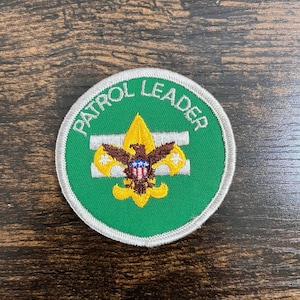 May include: A round green patch with the words "PATROL LEADER" in white lettering. The patch features a gold fleur-de-lis emblem with a brown eagle and a blue and white shield. The patch is trimmed with a silver border.