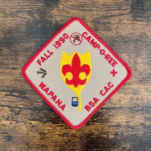 May include: A diamond-shaped patch with a red border and tan background. The patch features text that reads "FALL 1990 CAMP-O-REE WAPAH BSA CAC". A yellow and red fleur-de-lis design is in the center.