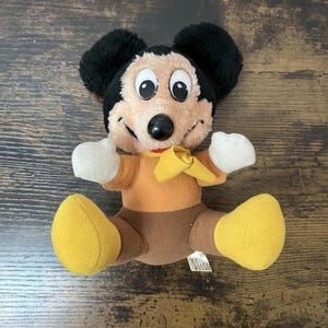 Puede incluir: Un peluche vintage de Mickey Mouse. El peluche presenta una cabeza negra con orejas grandes, una cara beige y una nariz negra. Lleva una camisa naranja, pantalones marrones, zapatos amarillos y una pajarita amarilla. El juguete está en posición sentada.