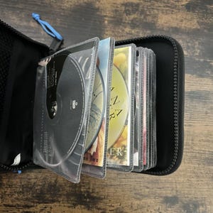 Puede incluir: Estuche negro para CD con cremallera, que contiene varios CD en fundas de plástico transparente. Los CD muestran carátulas de álbumes y texto. El estuche está abierto, revelando los CD. El fondo es una superficie de madera.