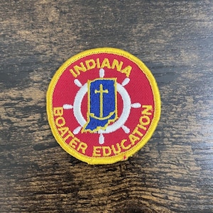 Pode incluir: Um patch redondo com as palavras "INDIANA BOATER EDUCATION" em letras amarelas sobre um fundo vermelho. O patch apresenta um design de volante branco e um emblema azul e branco com uma cruz.