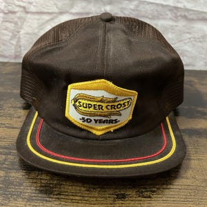 May include: A brown trucker cap with a mesh back and a curved peak. The front has a patch with the words "SUPER CROST" and "50 YEARS" in gold and white. The peak is trimmed with red and yellow stripes.