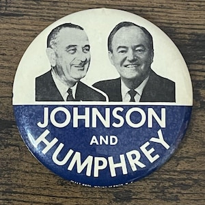 Puede incluir: Un botón de campaña vintage con retratos en blanco y negro de dos hombres sobre las palabras "JOHNSON AND HUMPHREY" en blanco sobre un fondo azul. El botón es redondo y tiene un alfiler en la parte posterior.