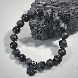 Könnte beinhalten: Ein schwarzes Perlenarmband mit einer Vielzahl von Formen und Texturen, darunter facettierte und glatte Perlen. Das Armband hat einen kleinen, runden Anhänger mit einem Unendlichkeitssymbol. Das Armband wird auf einer hellgrauen Oberfläche mit einem schwarzen Stein im Hintergrund präsentiert.