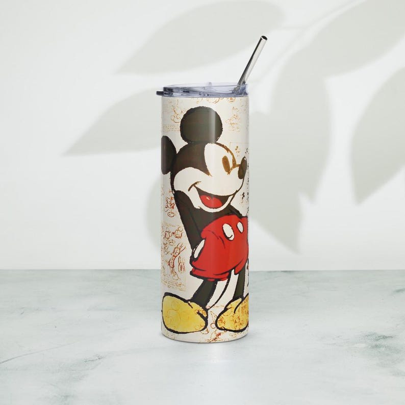 May include: A cylindrical stainless steel tumbler featuring a vintage-style illustration of Mickey Mouse wearing red shorts and yellow shoes. The background has a cream-colored parchment texture with faint sketches. A clear lid and metal straw are included.