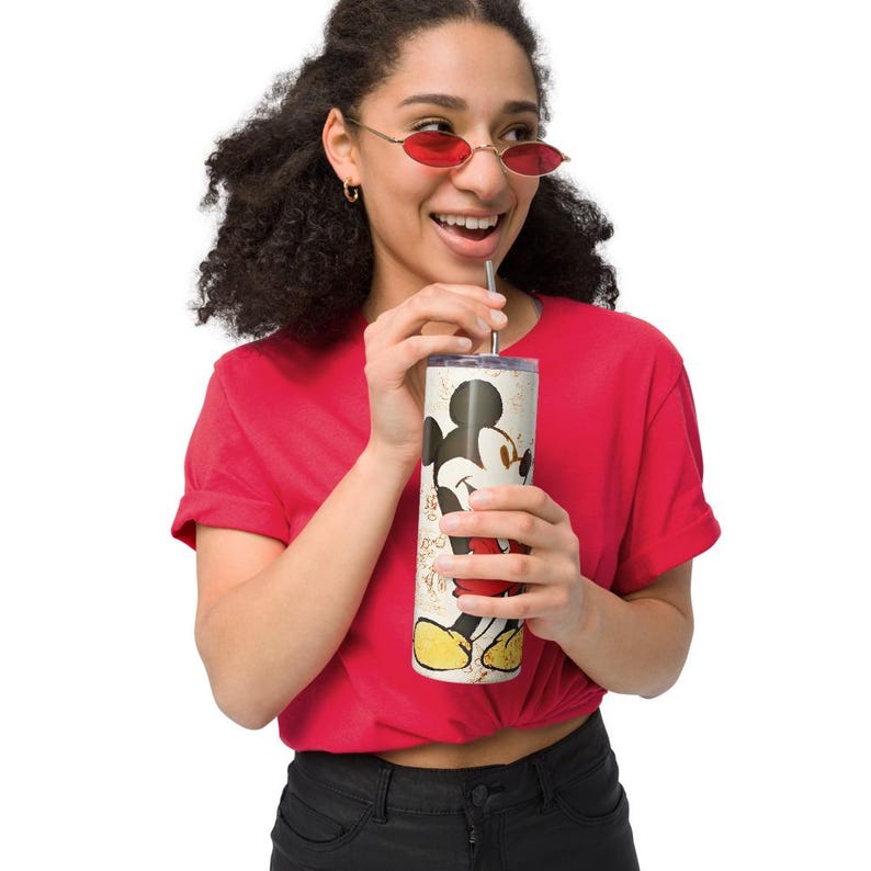 May include: A cylindrical stainless steel tumbler featuring a graphic illustration of Mickey Mouse with a red heart, set against a cream-colored background with subtle gold-toned accents and a metal straw inserted through the lid.