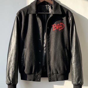 Op de afbeelding: Zwarte varsityjas met leren mouwen en het woord "BAD" in rood geborduurd op de borst. De jas heeft een knoopsluiting, een kraag en geribde manchetten. De jas hangt aan een houten kleerhanger.