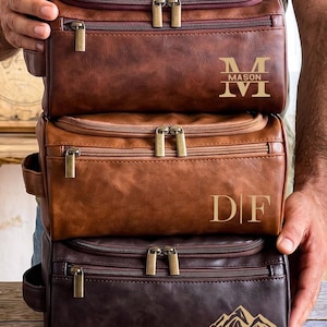 May include: Three rectangular toiletry bags stacked vertically in dark brown, medium brown, and deep espresso brown. Each bag features a top zip, a front zipped pocket, and antique brass hardware. Custom engravings include MASON, DF, and ROBERT.