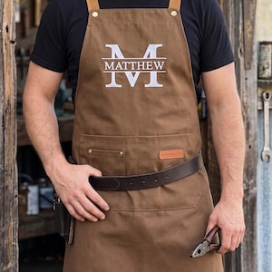 May include: A brown canvas apron featuring tan leather shoulder straps with brass rivets and a dark brown leather waist belt. The chest area displays the white text MATTHEW centred between two horizontal lines and a large letter M.
