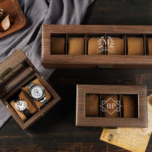Puede incluir: Cajas de reloj de madera con tapas de cristal e iniciales personalizadas. Una caja está abierta, mostrando dos relojes. Las otras cajas están cerradas, con cojines para guardar relojes. Las cajas están sobre una superficie de madera oscura.