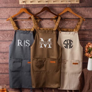 May include: Three aprons hanging on wooden hangers. The left is dark grey with white 'RS' embroidery. The centre is brown with white 'MATTHEW' embroidery. The right is beige with a black circular 'SRE' monogram. Each features two front pockets.