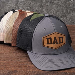 May include: A collection of trucker hats in various colors, including gray, black, and tan. Each hat features a mesh back and a front panel with a leather patch that reads "DAD". The hats are displayed on a wooden surface.