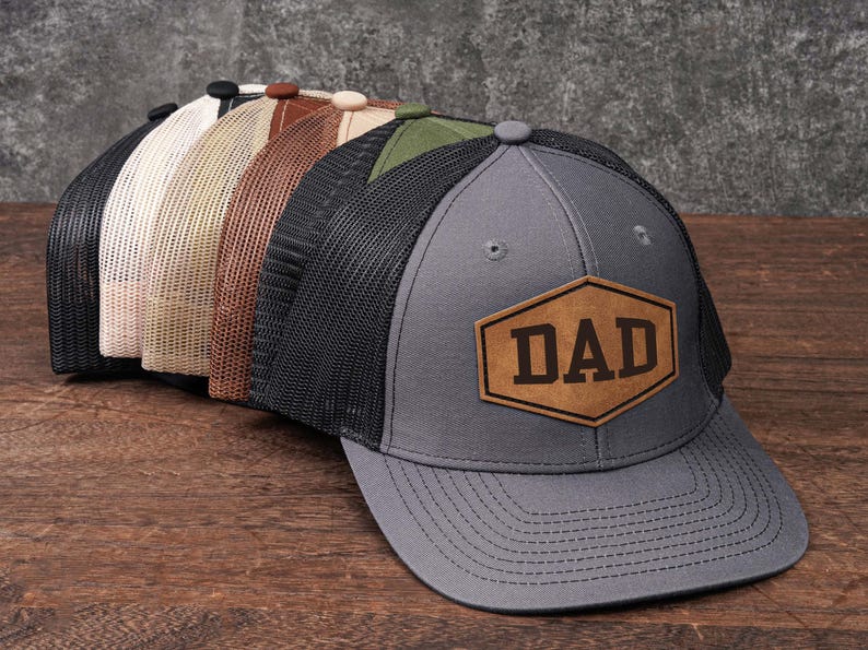 May include: A collection of trucker hats in various colors, including gray, black, and tan. Each hat features a mesh back and a front panel with a leather patch that reads "DAD". The hats are displayed on a wooden surface.