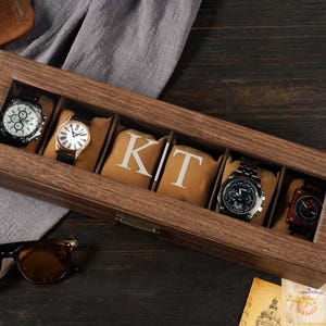 Peut inclure: Un coffret en bois pour montres avec un couvercle transparent, contenant cinq montres. Le coffret comporte des compartiments individuels avec des coussins beiges. Les lettres "KT" sont imprimées sur deux des coussins. Des lunettes de soleil sont au premier plan.