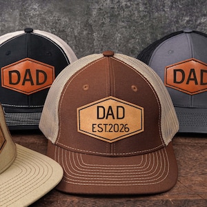 May include: Four trucker hats with the word "DAD" on a leather patch. The hats are in various colors: black, gray, brown, and tan. The brown hat has "EST.2026" below "DAD". Each hat has a mesh back and a curved brim.