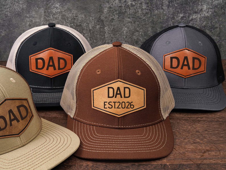 May include: Four trucker hats with the word "DAD" on a leather patch. The hats are in various colors: black, gray, brown, and tan. The brown hat has "EST.2026" below "DAD". Each hat has a mesh back and a curved brim.