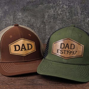 May include: Two baseball caps with a mesh back and a leather patch. One is brown with "DAD" in a hexagon, the other is green with "DAD EST.1997". Both are on a wooden surface.