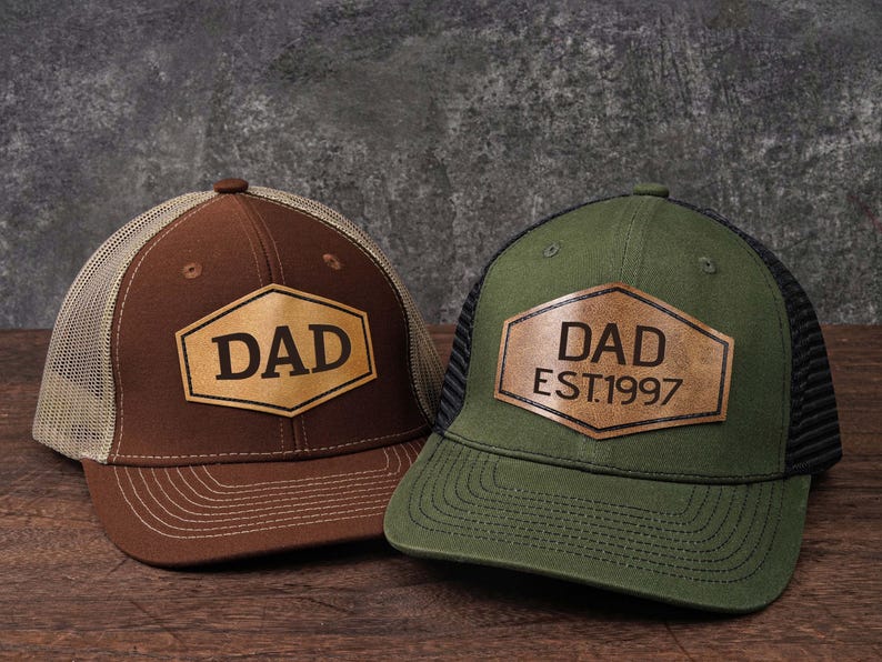 May include: Two baseball caps with a mesh back and a leather patch. One is brown with "DAD" in a hexagon, the other is green with "DAD EST.1997". Both are on a wooden surface.