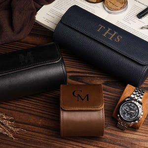 Pu&ograve; includere: Un assortimento di custodie per orologi personalizzate in pelle nera, blu navy e marrone. Le custodie sono goffrate con iniziali. Un orologio da polso argentato con quadrante nero &egrave; appoggiato su un cuscino marrone. Su una superficie di legno.