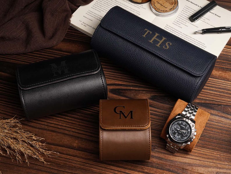 Pu&ograve; includere: Un assortimento di custodie per orologi personalizzate in pelle nera, blu navy e marrone. Le custodie sono goffrate con iniziali. Un orologio da polso argentato con quadrante nero &egrave; appoggiato su un cuscino marrone. Su una superficie di legno.