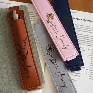 Puede incluir: Estuches de cuero para bolígrafos en marrón, rosa, gris y azul marino. Cada uno tiene un diseño floral y un nombre: Lucas, Emily y Henry. Un bolígrafo está en el estuche marrón. Los estuches están sobre un libro.