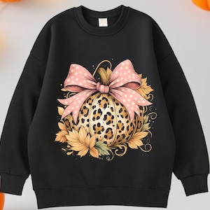 May include: Black sweatshirt featuring a pumpkin design. The pumpkin has a leopard print pattern and is adorned with a pink bow with white polka dots. Autumn leaves surround the pumpkin, creating a festive fall theme.