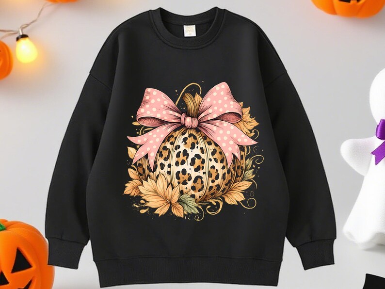 May include: Black sweatshirt featuring a pumpkin design. The pumpkin has a leopard print pattern and is adorned with a pink bow with white polka dots. Autumn leaves surround the pumpkin, creating a festive fall theme.