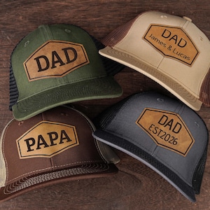 May include: Four trucker hats with leather patches. One is olive green with "DAD", another is tan with "DAD James & Lucas", a third is brown with "PAPA", and the last is gray with "DAD EST2026". All have mesh backs.
