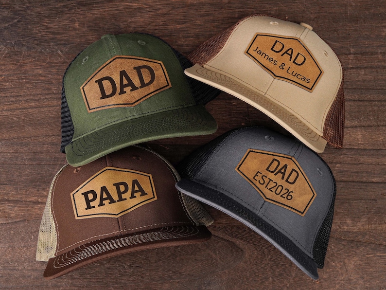 May include: Four trucker hats with leather patches. One is olive green with "DAD", another is tan with "DAD James & Lucas", a third is brown with "PAPA", and the last is gray with "DAD EST2026". All have mesh backs.