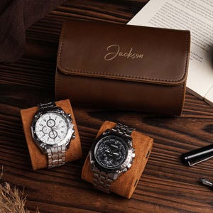 Pu&ograve; includere: Un astuccio per orologi in pelle marrone con il nome "Jackson" impresso sul davanti. Due orologi argentati con quadranti bianchi e neri poggiano su cuscini marroni. Sono visibili anche una penna, una candela e un libro aperto.