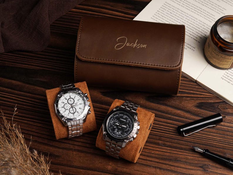 Pu&ograve; includere: Un astuccio per orologi in pelle marrone con il nome "Jackson" impresso sul davanti. Due orologi argentati con quadranti bianchi e neri poggiano su cuscini marroni. Sono visibili anche una penna, una candela e un libro aperto.