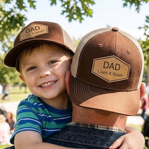 Peut inclure: Deux casquettes de camionneur marron avec le mot "DAD" et les noms "Liam & Noah" sur un patch en cuir. L'un des chapeaux est porté par un enfant, l'autre par un adulte. Les chapeaux ont un devant marron et un dos en filet beige. L'arrière-plan est un cadre de parc.