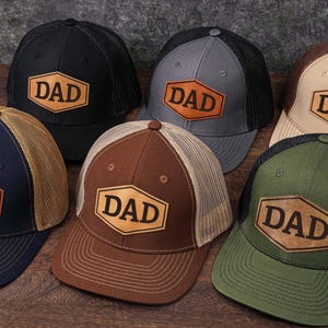 May include: A collection of six trucker hats in various colors, including black, navy, gray, brown, and green. Each hat features a leather patch with the word "DAD" in a bold font. The hats have mesh backs and curved brims.