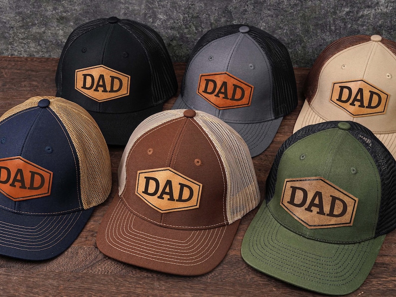 May include: A collection of six trucker hats in various colors, including black, navy, gray, brown, and green. Each hat features a leather patch with the word "DAD" in a bold font. The hats have mesh backs and curved brims.