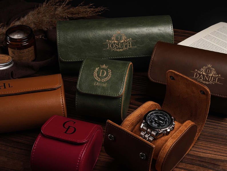 Pu&ograve; includere: Un assortimento di custodie per orologi in pelle in vari colori, tra cui verde, marrone e rosso. Alcune custodie sono personalizzate con nomi e iniziali. Un orologio argentato con quadrante nero &egrave; esposto in una custodia marrone aperta.