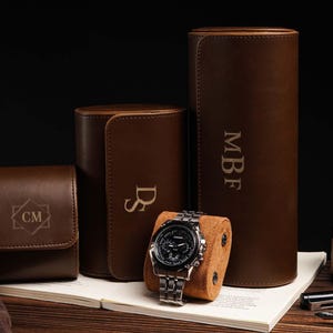 Pu&ograve; includere: Una collezione di custodie per orologi in pelle marrone e un orologio. Le custodie sono cilindriche, rettangolari e quadrate, con iniziali incise. Un orologio argentato con quadrante nero &egrave; appoggiato su un cuscino marrone.
