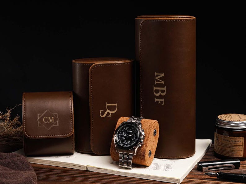 Pu&ograve; includere: Una collezione di custodie per orologi in pelle marrone e un orologio. Le custodie sono cilindriche, rettangolari e quadrate, con iniziali incise. Un orologio argentato con quadrante nero &egrave; appoggiato su un cuscino marrone.