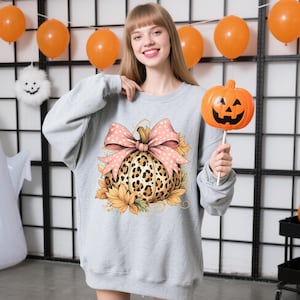 May include: A light gray sweatshirt featuring a pumpkin design with a leopard print pattern, a pink bow, and sunflowers. The person is holding an orange pumpkin on a stick. Halloween decorations are in the background.