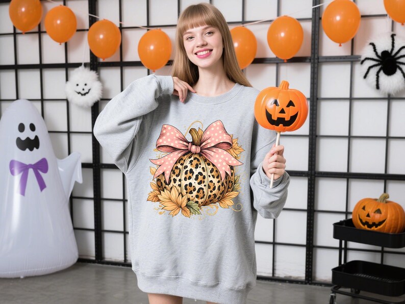 May include: A light gray sweatshirt featuring a pumpkin design with a leopard print pattern, a pink bow, and sunflowers. The person is holding an orange pumpkin on a stick. Halloween decorations are in the background.