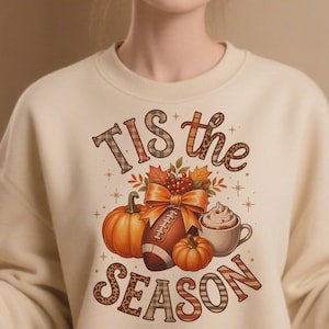 K&ouml;nnte beinhalten: Cremefarbenes Sweatshirt mit einem herbstlichen Motiv. Das Design umfasst K&uuml;rbisse, einen Football, Herbstbl&auml;tter und eine Tasse Kaffee mit Schlagsahne. Der Text "Tis the Season" wird in verschiedenen Schriftarten und Mustern angezeigt.