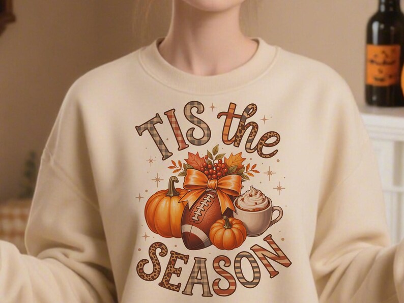 K&ouml;nnte beinhalten: Cremefarbenes Sweatshirt mit einem herbstlichen Motiv. Das Design umfasst K&uuml;rbisse, einen Football, Herbstbl&auml;tter und eine Tasse Kaffee mit Schlagsahne. Der Text "Tis the Season" wird in verschiedenen Schriftarten und Mustern angezeigt.
