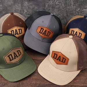 May include: Five baseball caps with mesh backs and curved brims are displayed. Each hat features a leather patch with the word "DAD" in a bold font. Colors include brown, green, gray, navy, and tan.