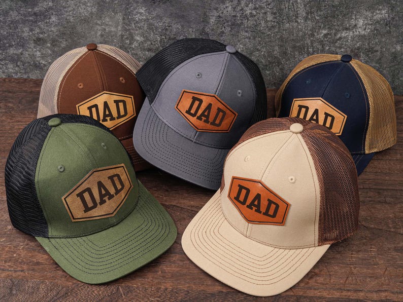 May include: Five baseball caps with mesh backs and curved brims are displayed. Each hat features a leather patch with the word "DAD" in a bold font. Colors include brown, green, gray, navy, and tan.