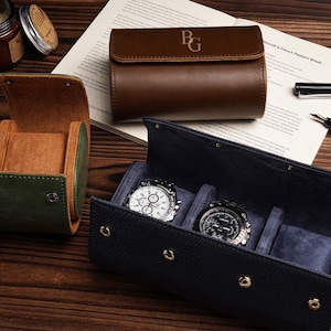 Pu&ograve; includere: Custodie per orologi in pelle verde, marrone e blu navy. Due orologi con quadranti argentati e neri sono esposti in una custodia blu navy aperta. &Egrave; presente anche una custodia in pelle marrone con iniziali.