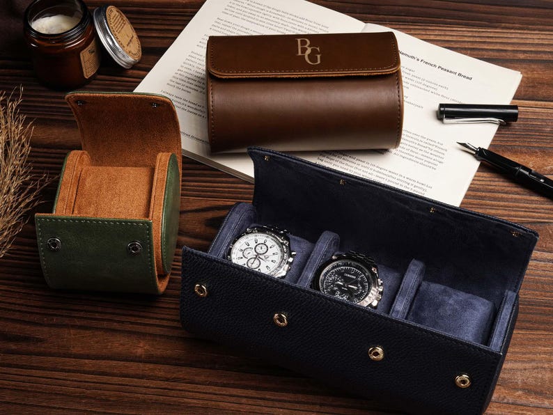 Pu&ograve; includere: Custodie per orologi in pelle verde, marrone e blu navy. Due orologi con quadranti argentati e neri sono esposti in una custodia blu navy aperta. &Egrave; presente anche una custodia in pelle marrone con iniziali.